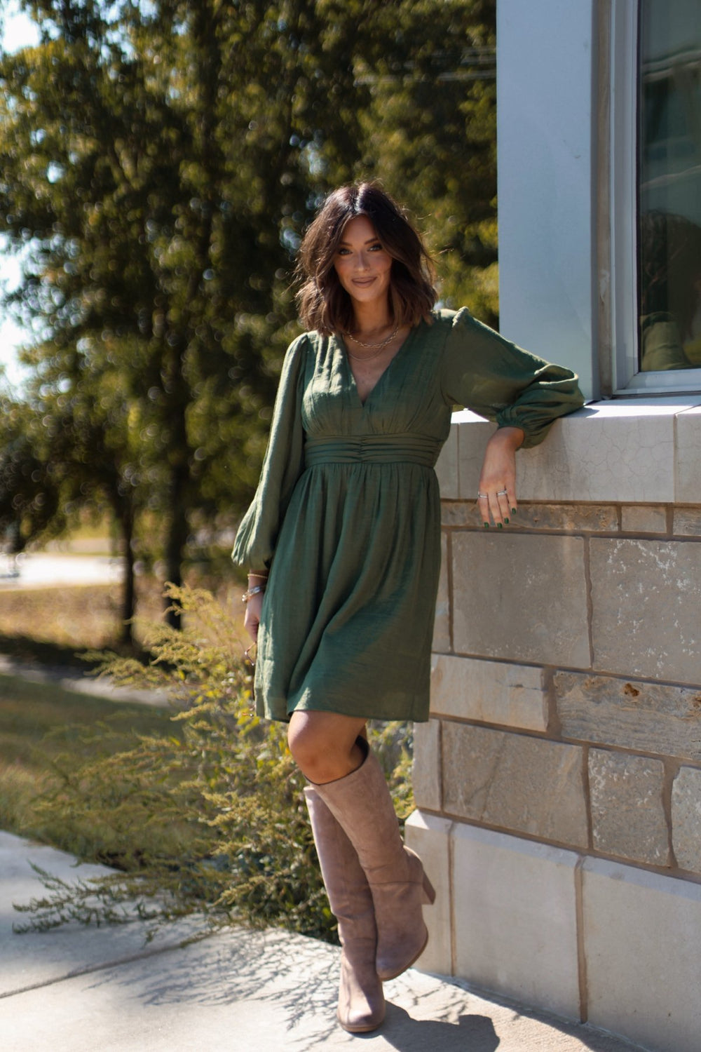 Woman in the Vanessa Green Ruched Mini Dress - FINAL SALE and knee-high boots leans against a sunlit stone wall outdoors.