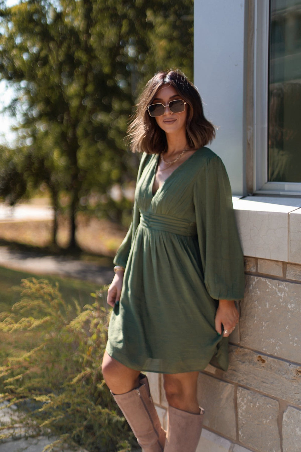 Woman in the Vanessa Green Ruched Mini Dress - FINAL SALE poses by a stone wall, with sunlight and greenery highlighting the scene.