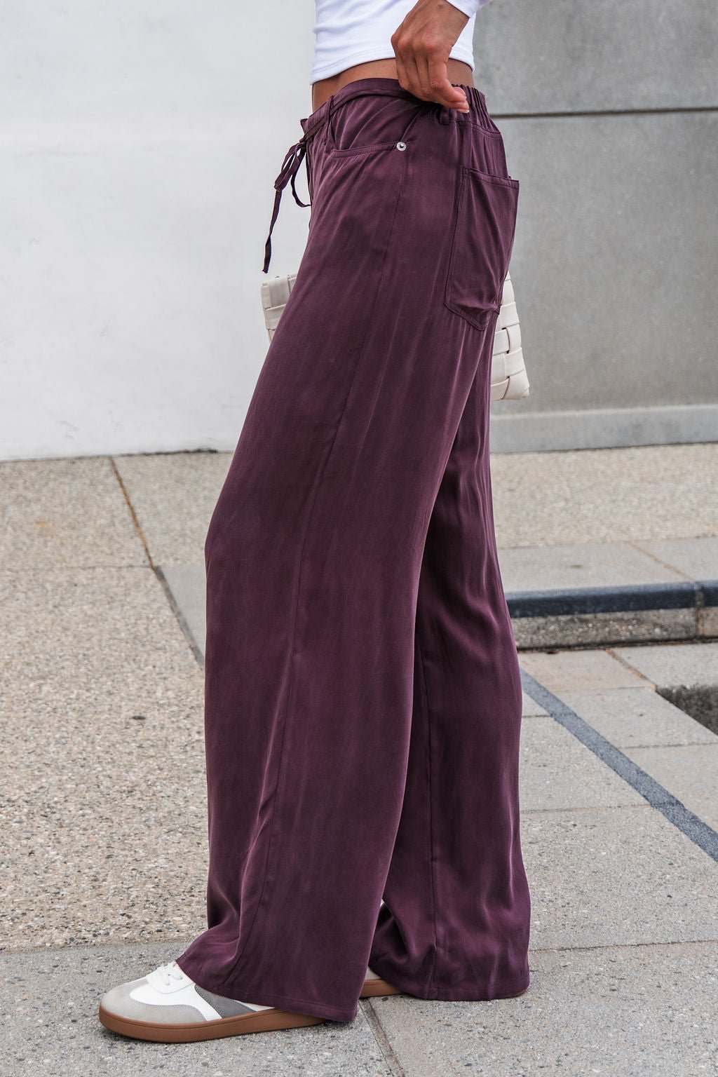 Person in brown Vera Wide Leg Pants with an elastic waistband, white sneakers, and a woven clutch stands on a sidewalk.
