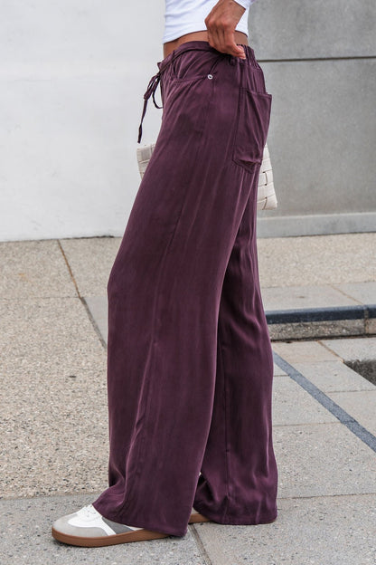 Person in brown Vera Wide Leg Pants with an elastic waistband, white sneakers, and a woven clutch stands on a sidewalk.