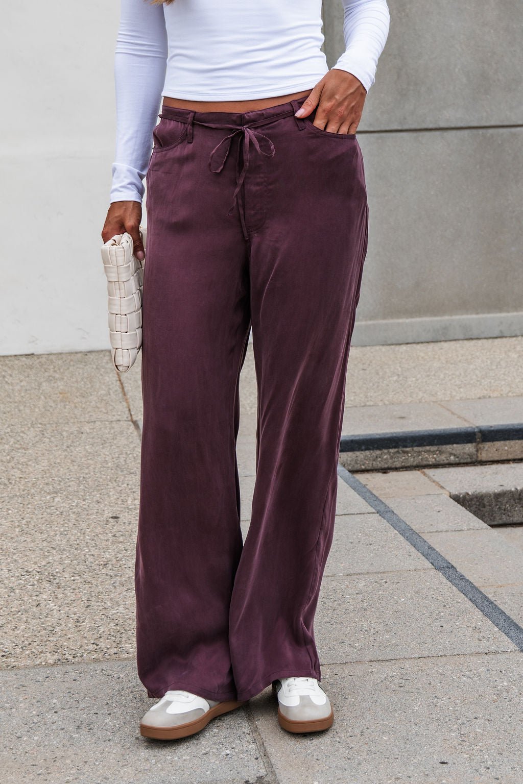 Woman wearing loose brown Vera Wide Leg Pants with an elastic waist, white top, sneakers, and holding a white woven clutch on the sidewalk.