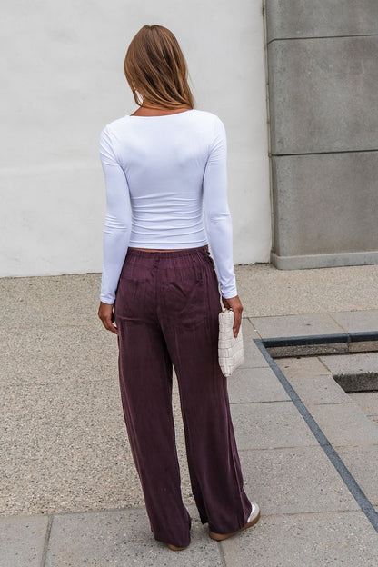 Woman stands outdoors in a white top and Vera Wide Leg Pants - Brown, featuring an elastic waistband, holding a white clutch.