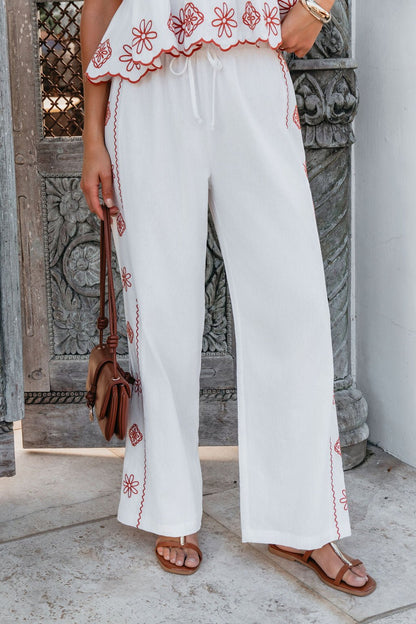 Woman in Veranda White Embroidered Pants and sandals, holding a brown purse, stands by a carved wooden door and column.