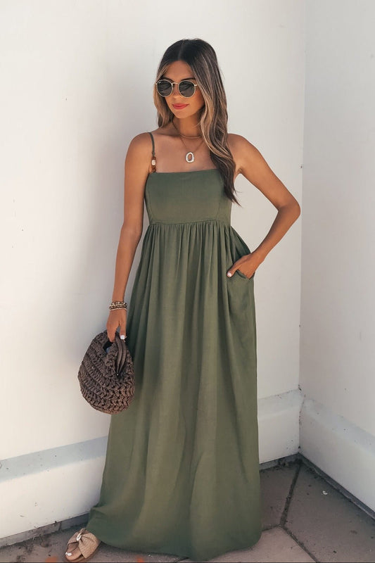 Woman in Verdant Olive Smocked Linen Maxi Dress with sunglasses, sandals, and woven bag, standing by a white wall.