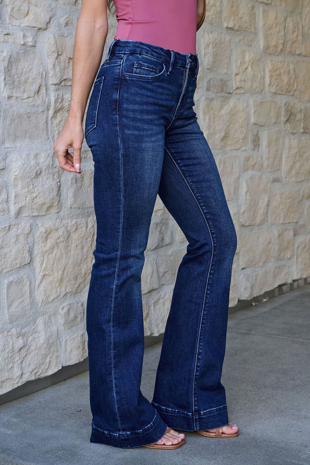 A woman in Vervet Dark Wash Mid Rise Flare Jeans, a pink sleeveless top, and sandals stands by a stone wall.