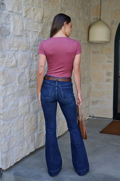 A woman faces away in a pink top and Vervet Dark Wash Mid Rise Flare Jeans | Pre Order, holding a brown bag at her side.