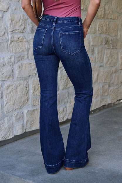 Someone in Vervet Dark Wash Mid Rise Flare Jeans stands on a sidewalk near a stone wall, facing away from the camera.