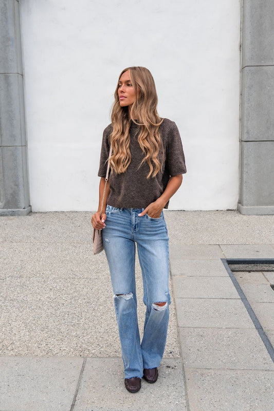Woman with long wavy hair wears Vervet Distressed Raw Hem Jeans - Light Wash, a dark top, and flats by a white and gray wall outdoors.