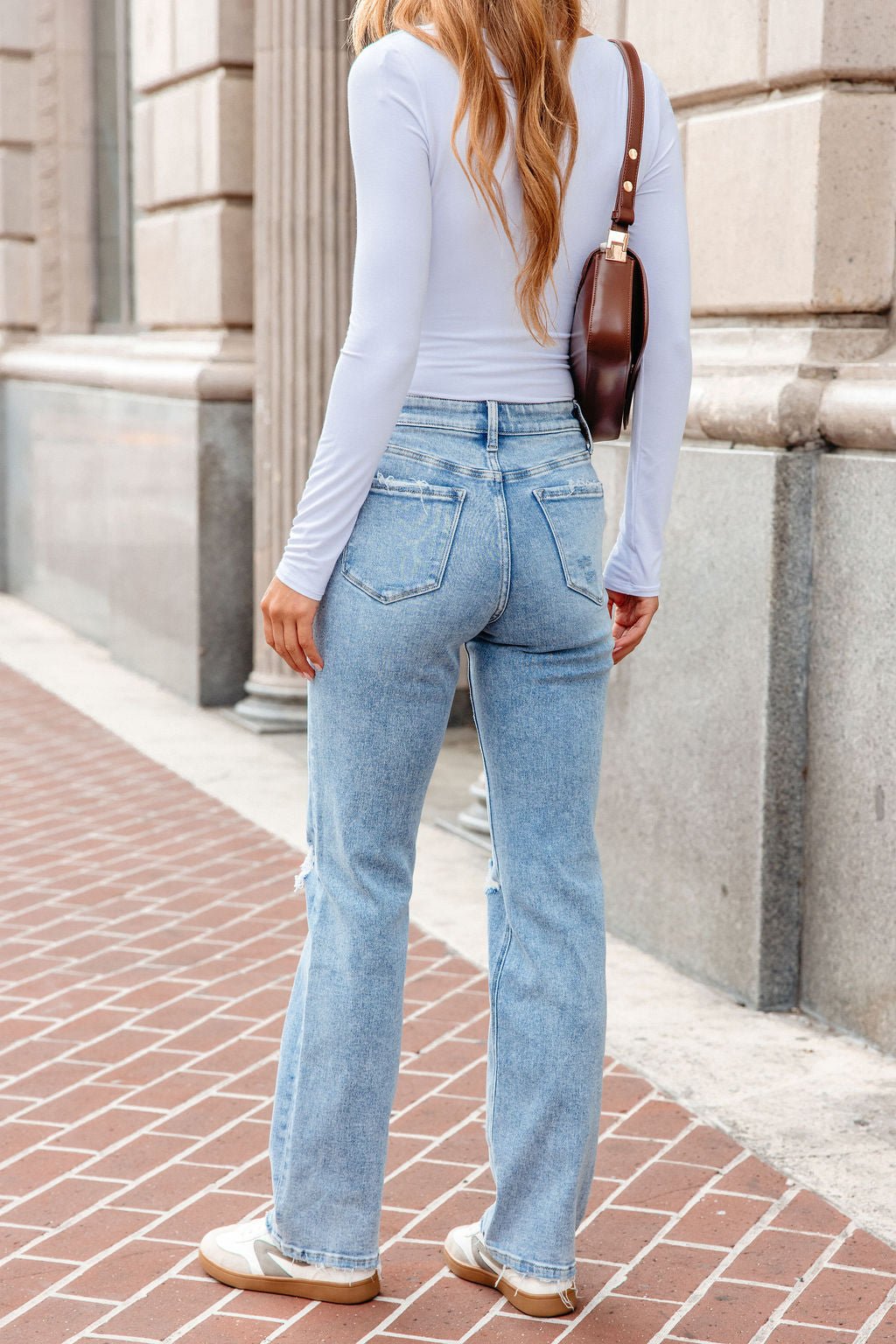 Woman in a white long-sleeve top, Vervet Distressed Straight Leg Jeans - Light Wash, sneakers, and a brown shoulder bag on a brick sidewalk.