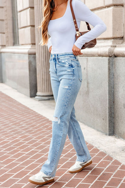 Woman in a white top, Vervet Distressed Straight Leg Jeans - Light Wash, and sneakers walks on a brick sidewalk carrying a brown bag.