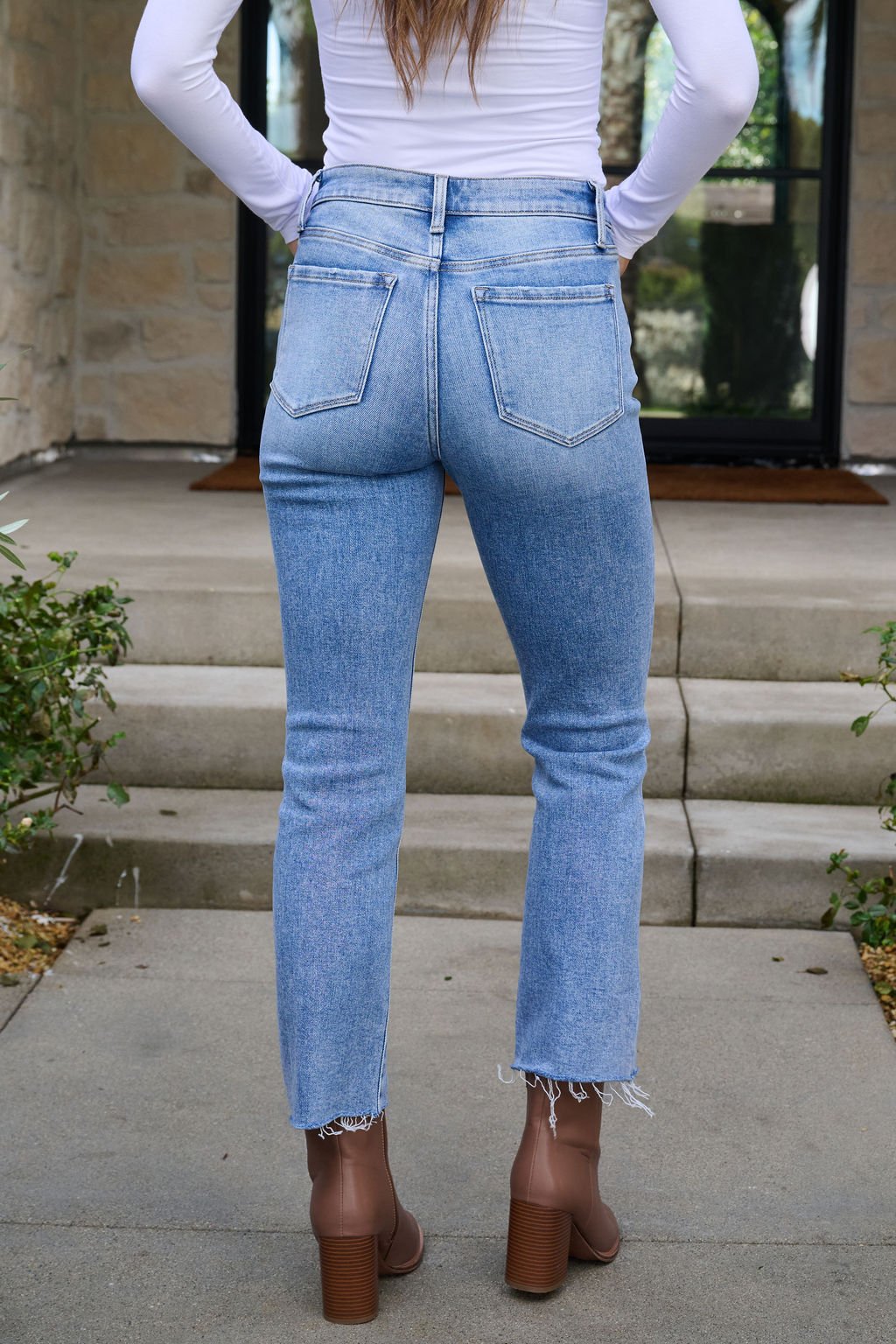 A woman in Vervet Light Wash Cropped Flare Jeans and brown boots stands on a path facing stone steps.