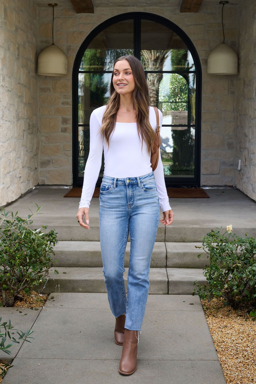 A woman in Vervet Light Wash Cropped Flare Jeans and a white top walks on a stone path toward arched double glass doors.