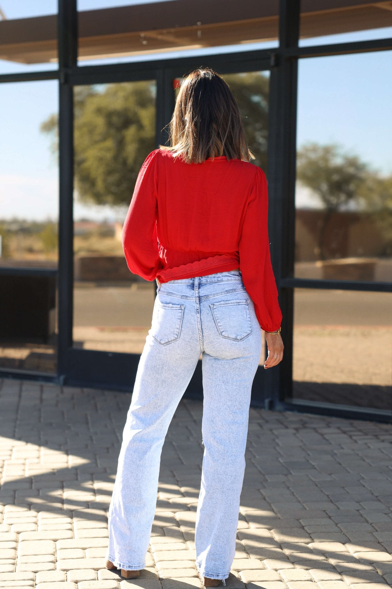 A woman in a red blouse and Vervet Light Wash Distressed Wide Leg Jeans stands outside on a paved surface, facing away.
