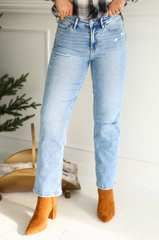Person wearing Vervet Light Wash High Rise Cropped Jeans and tan suede heeled boots, standing indoors near a pine tree and fireplace accessories.