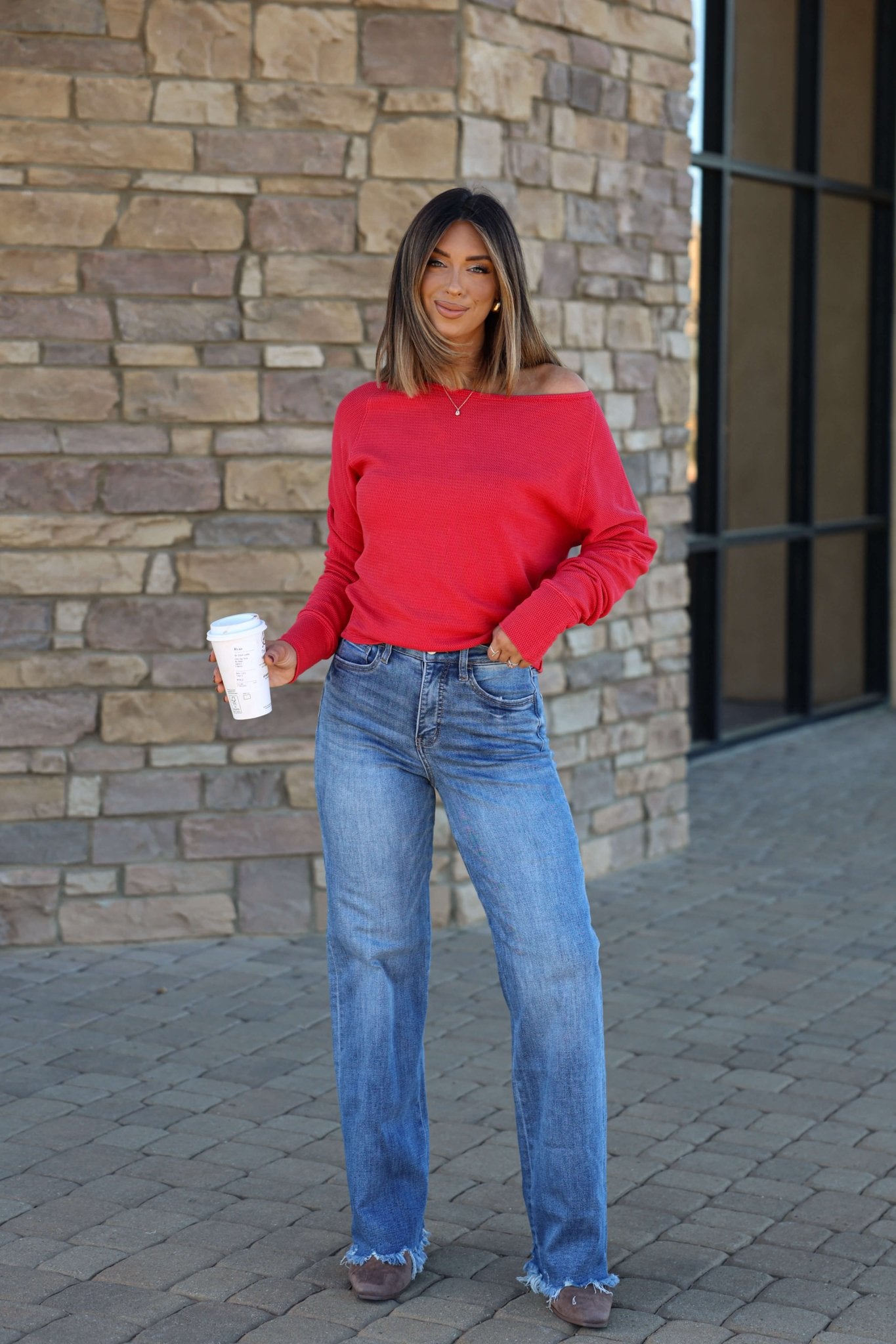 Woman in a red sweater and Vervet Medium Wash 90s Vintage Jeans holds a coffee cup, standing on stone pavement by a brick wall.