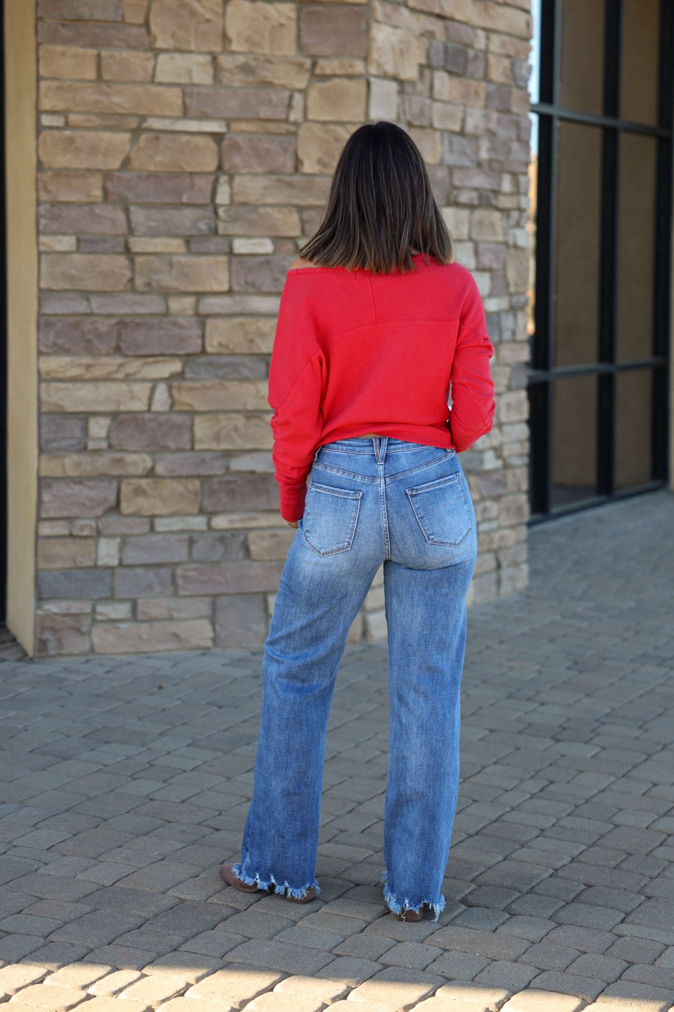 A woman in a red top and Vervet Medium Wash 90s Vintage Jeans with a distressed hem stands on stone pavement, facing a brick wall.