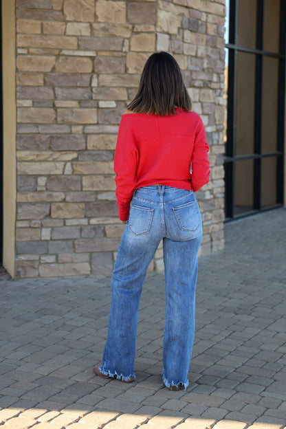 A woman in a red top and Vervet Medium Wash 90s Vintage Jeans with a distressed hem stands on stone pavement, facing a brick wall.