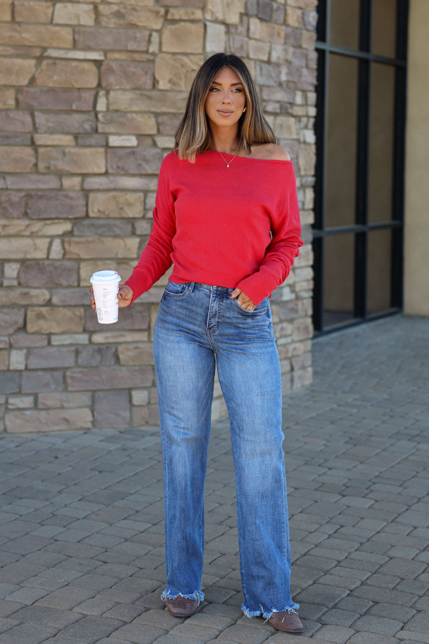 Woman in Vervet Medium Wash 90s Vintage Jeans, red off-shoulder top, holding coffee on stone pavement by a brick wall.