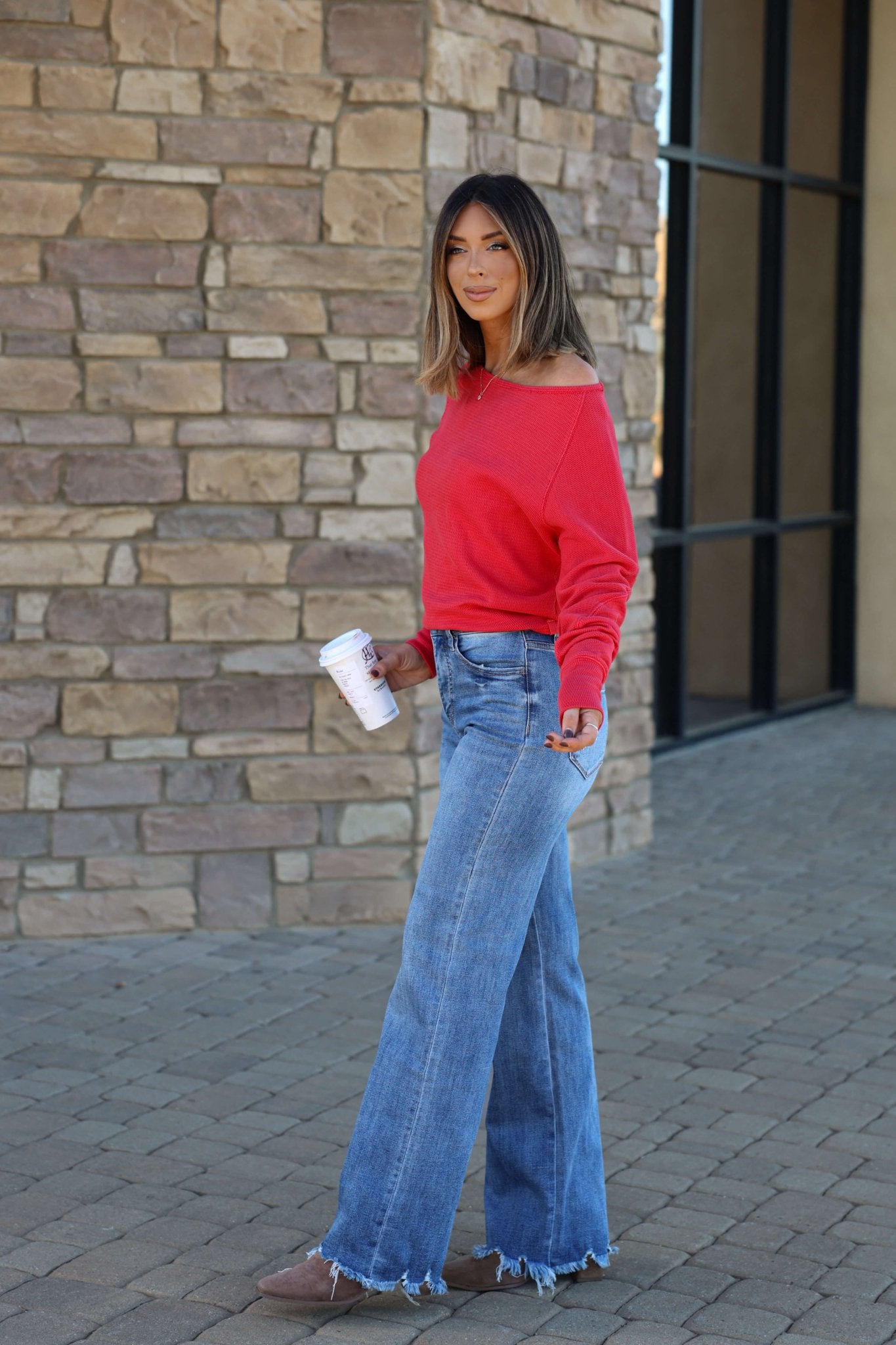 Woman in a red sweater wearing Vervet Medium Wash 90s Vintage Jeans with distressed hem, holding a coffee cup by a stone wall.