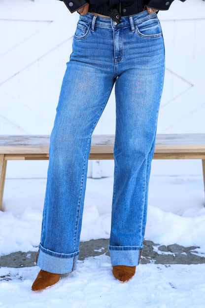 Person wearing Vervet Medium Wash High Rise Cuffed Jeans and brown shoes stands on snow in front of a wooden bench.
