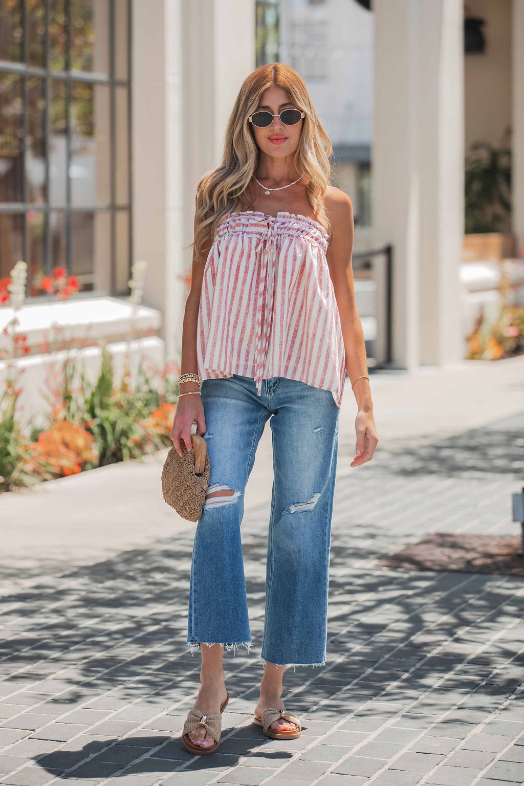 Woman in sunglasses wears a striped sleeveless top, Vervet Raw Hem Distressed Jeans - Medium Wash, sandals, and a woven bag outdoors.