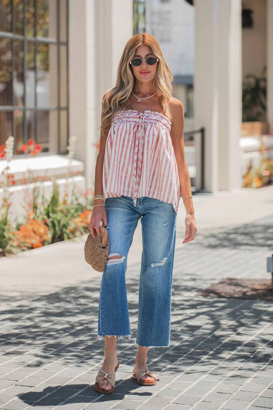Woman in sunglasses wears a striped sleeveless top, Vervet Raw Hem Distressed Jeans - Medium Wash, sandals, and a woven bag outdoors.
