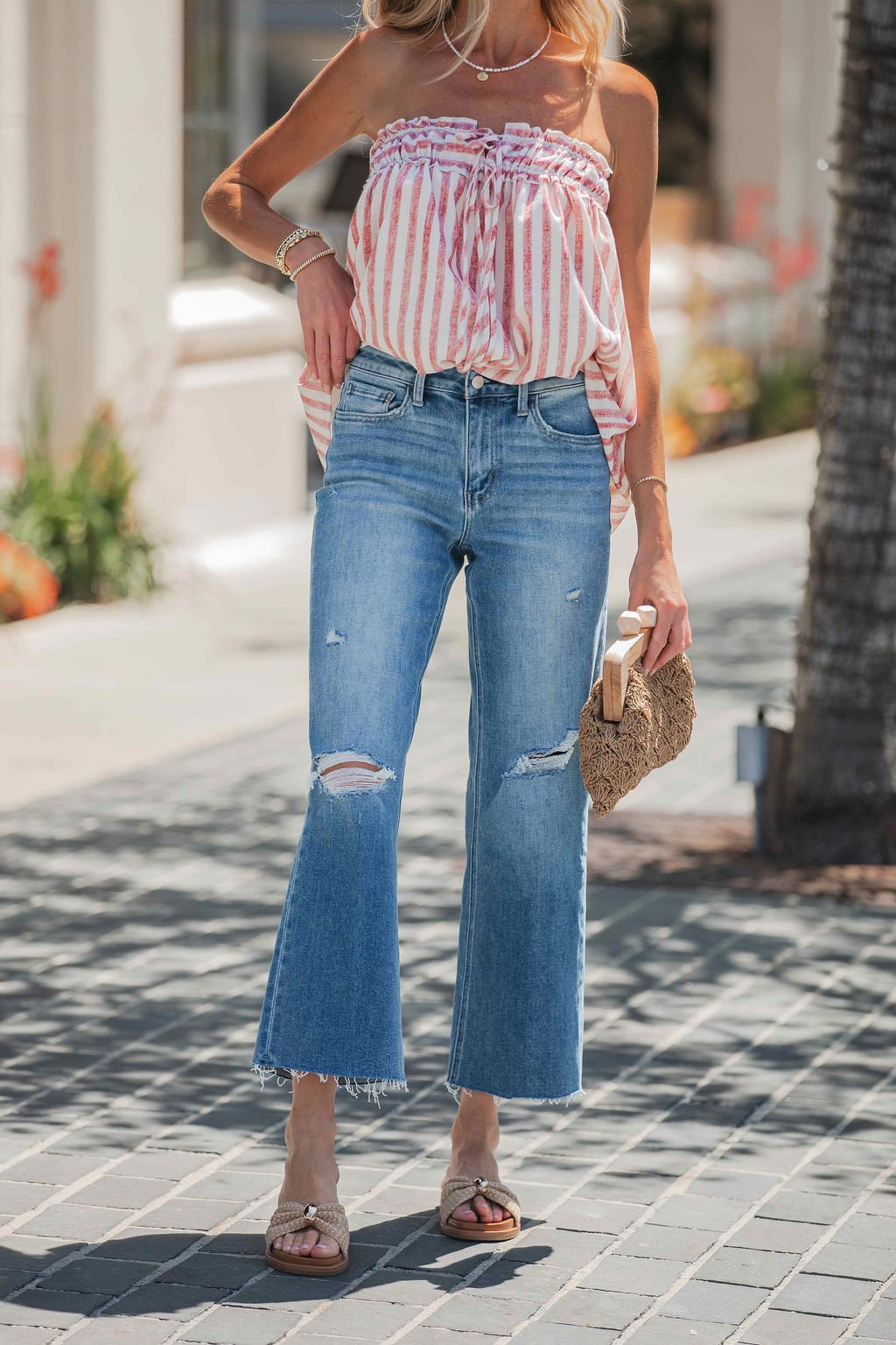Woman wearing Vervet Raw Hem Distressed Jeans - Medium Wash, pink striped top, sandals, and holding a woven clutch on a sunny street.