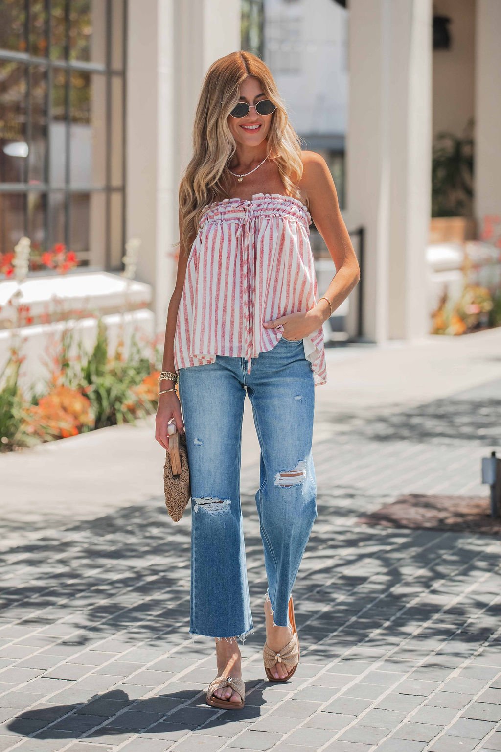 Woman in sunglasses, striped strapless top, Vervet Raw Hem Distressed Jeans - Medium Wash, sandals, holding a woven clutch outdoors.