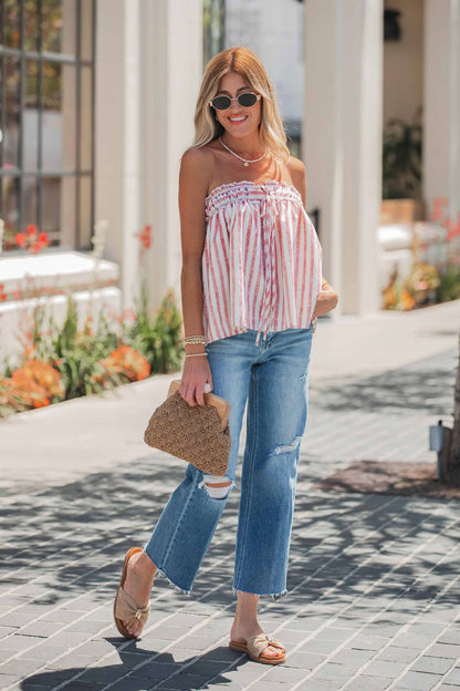 Woman in sunglasses, striped strapless top, Vervet Raw Hem Distressed Jeans - Medium Wash, sandals, and a woven handbag walks outside.