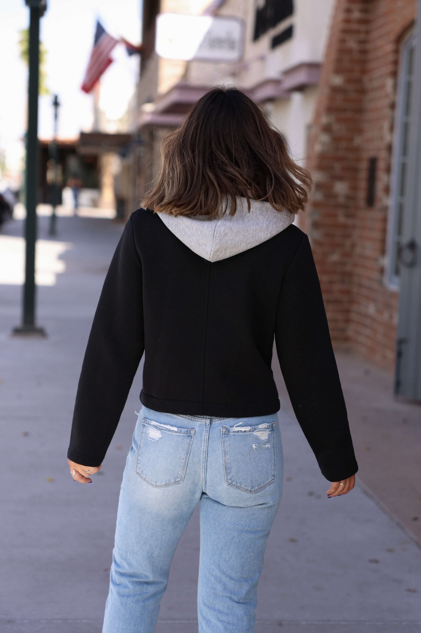 A person with shoulder-length brown hair walks on a sidewalk, wearing a Vintage Havana Black Contrast Hoodie Jacket.