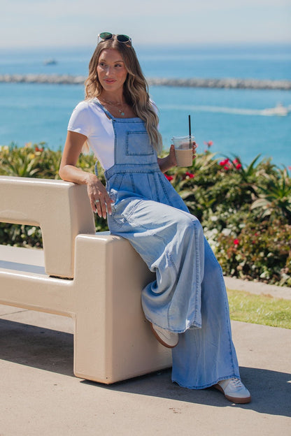 Woman in a Vintage Havana Denim Wide Leg Jumpsuit - FINAL SALE sits by the ocean, drink in hand, with flowers and water behind her.