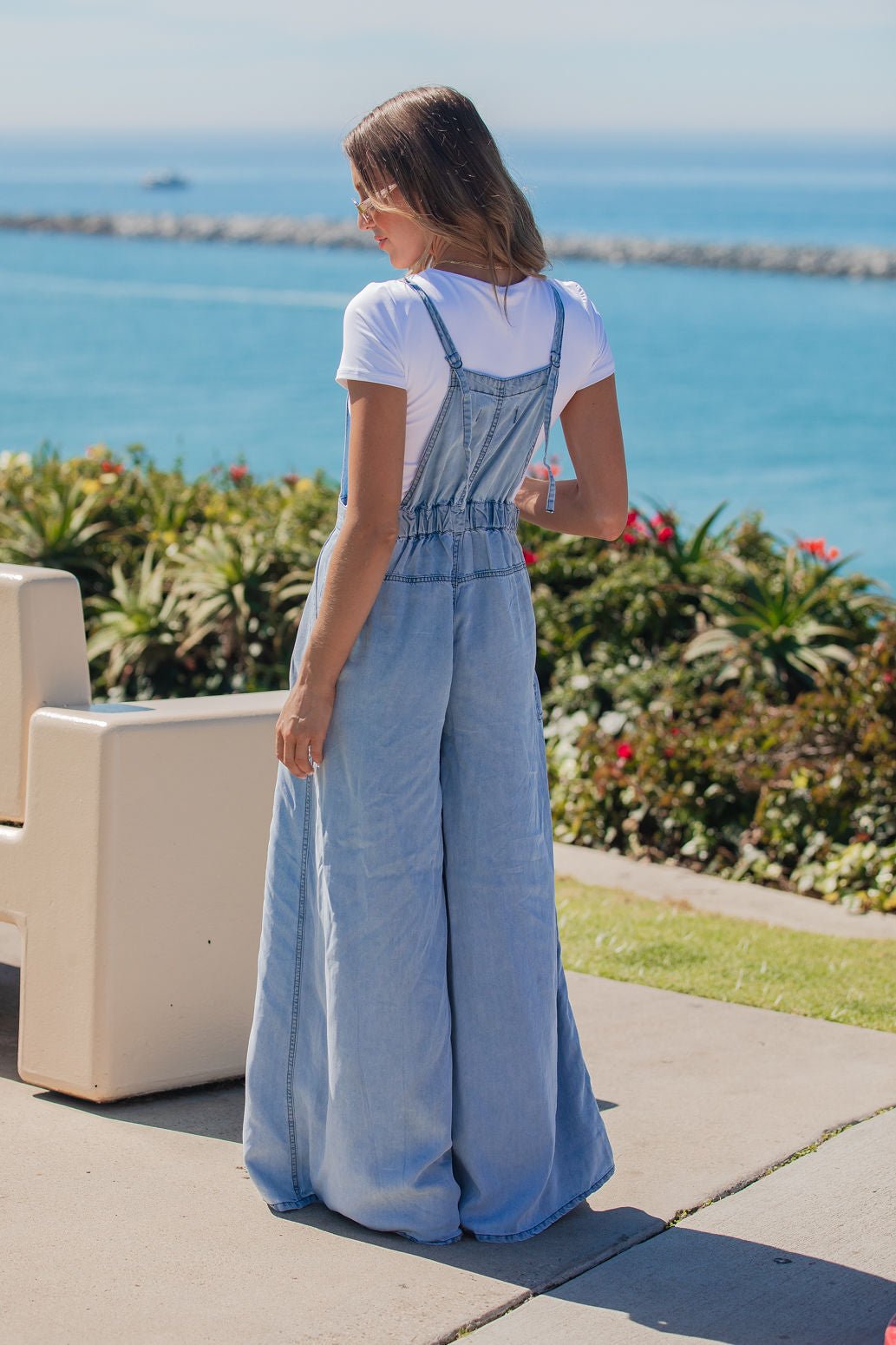 Woman in a Vintage Havana Denim Wide Leg Jumpsuit stands outdoors by the ocean, with plants and benches nearby.