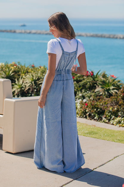 Woman in a Vintage Havana Denim Wide Leg Jumpsuit stands outdoors by the ocean, with plants and benches nearby.