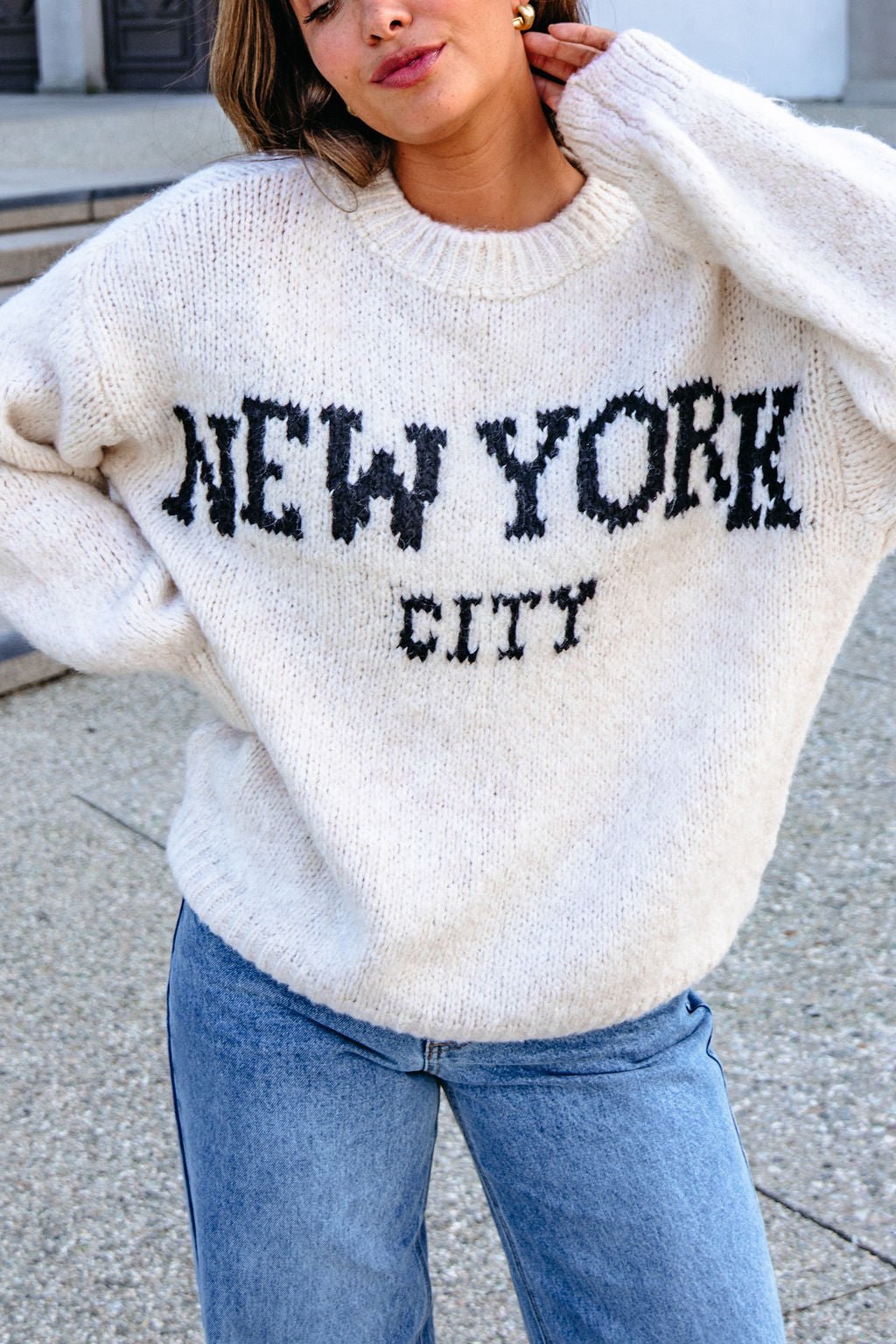 Person wears a Vintage Havana Ecru New York City Jacquard Sweater with blue jeans, standing outdoors.