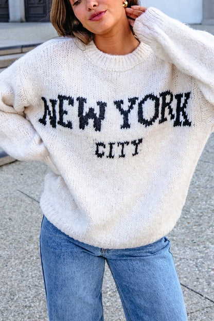 Person wears a Vintage Havana Ecru New York City Jacquard Sweater with blue jeans, standing outdoors.