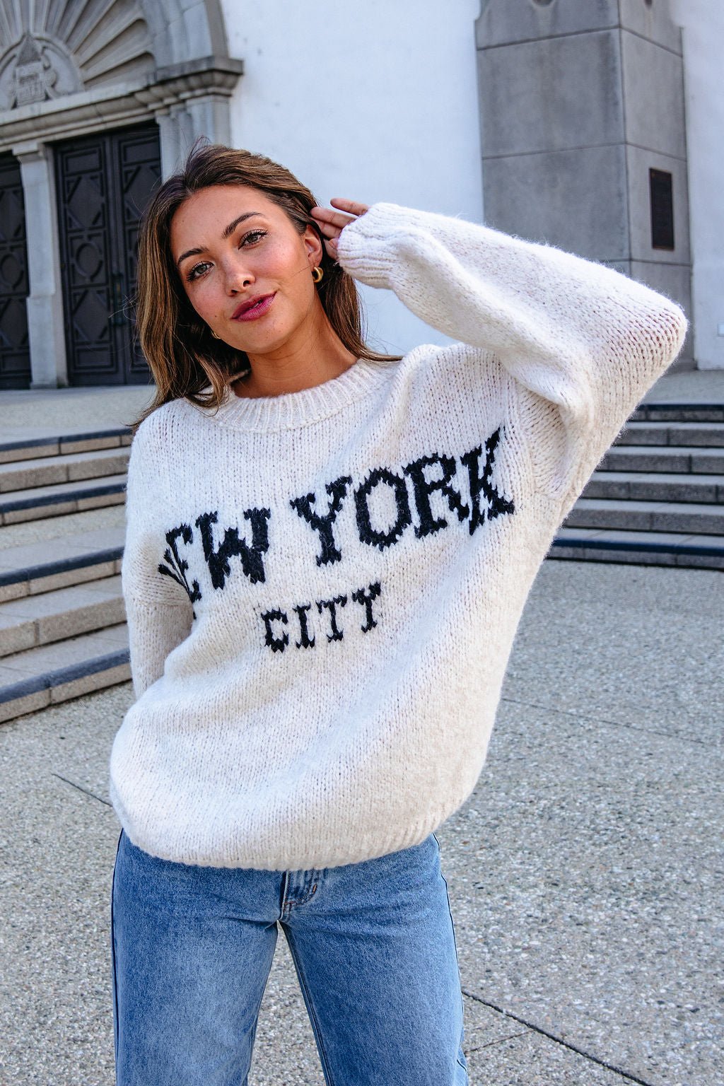 Woman in a Vintage Havana Ecru New York City Jacquard Sweater and blue jeans poses on stone steps, touching her hair and smiling.