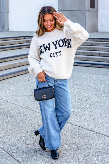 A woman in a Vintage Havana Ecru New York City Jacquard Sweater and wide-leg jeans smiles outdoors with a black bag and boots.