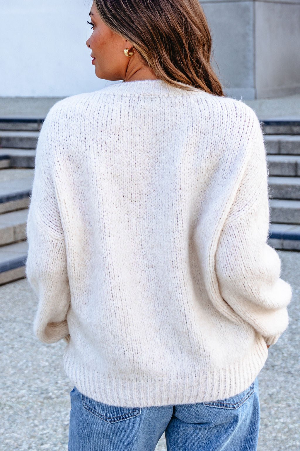 A woman in a Vintage Havana Ecru New York City Jacquard Sweater and blue jeans stands outdoors, her long brown hair flowing behind her.