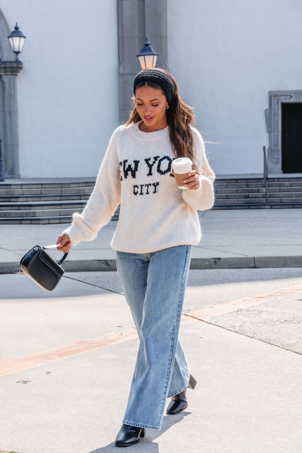 Woman in Vintage Havana Ecru New York City Jacquard Sweater, jeans, and boots, walking outside with coffee and a black handbag.