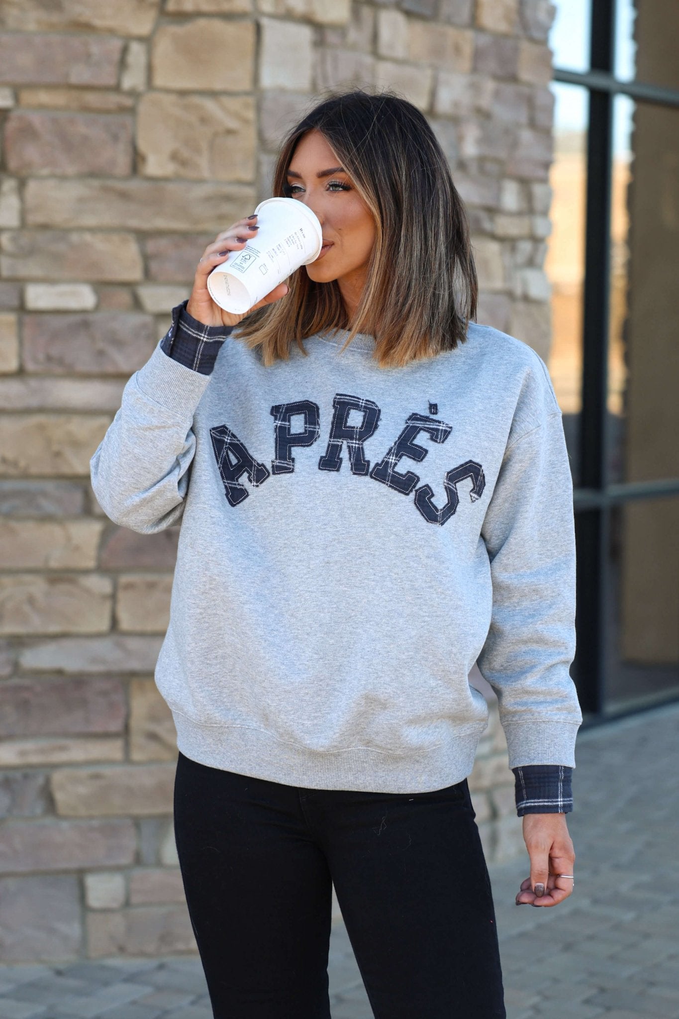 A woman with shoulder-length hair wears a Vintage Havana Grey Apres French Terry Sweatshirt, sipping from a white cup by a brick wall.