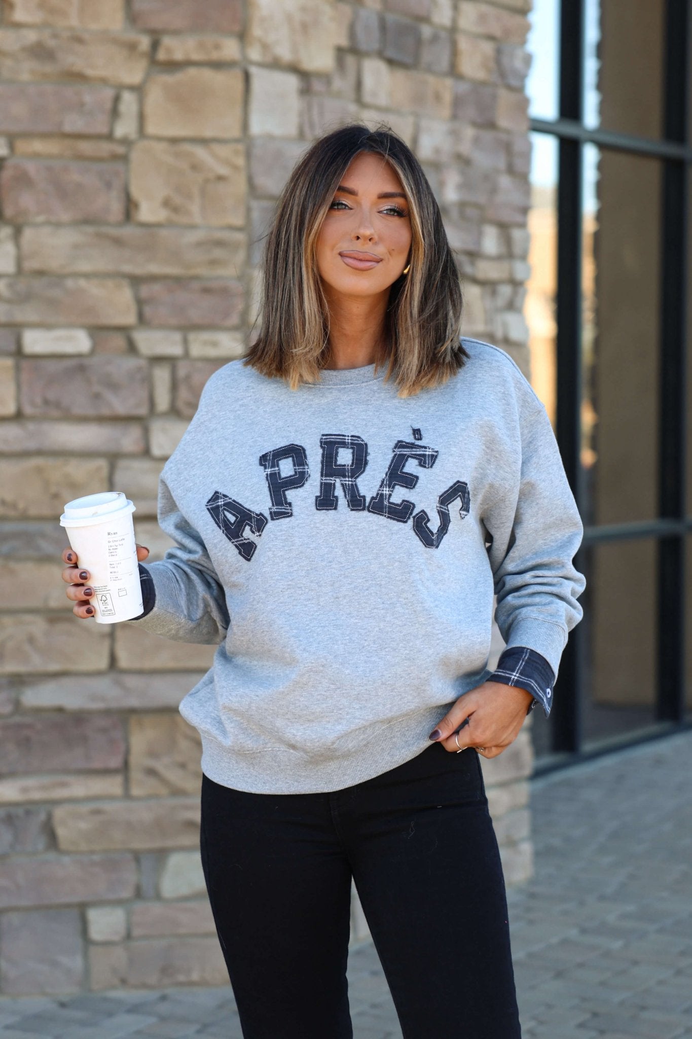 Woman with shoulder-length hair in a Vintage Havana Grey Apres French Terry Sweatshirt and black pants holds coffee by a stone wall.