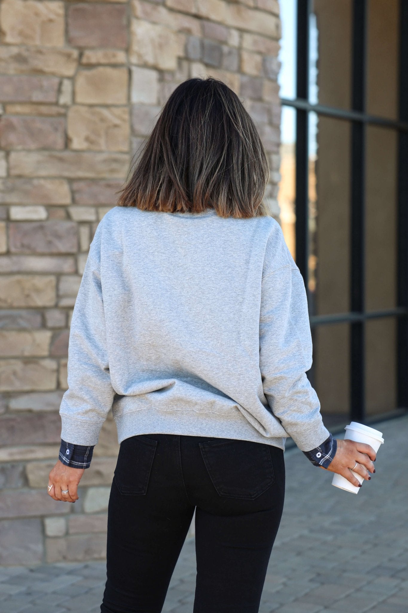 A woman with shoulder-length hair wears a Vintage Havana Grey Apres French Terry Sweatshirt and black pants, holding a white coffee cup.