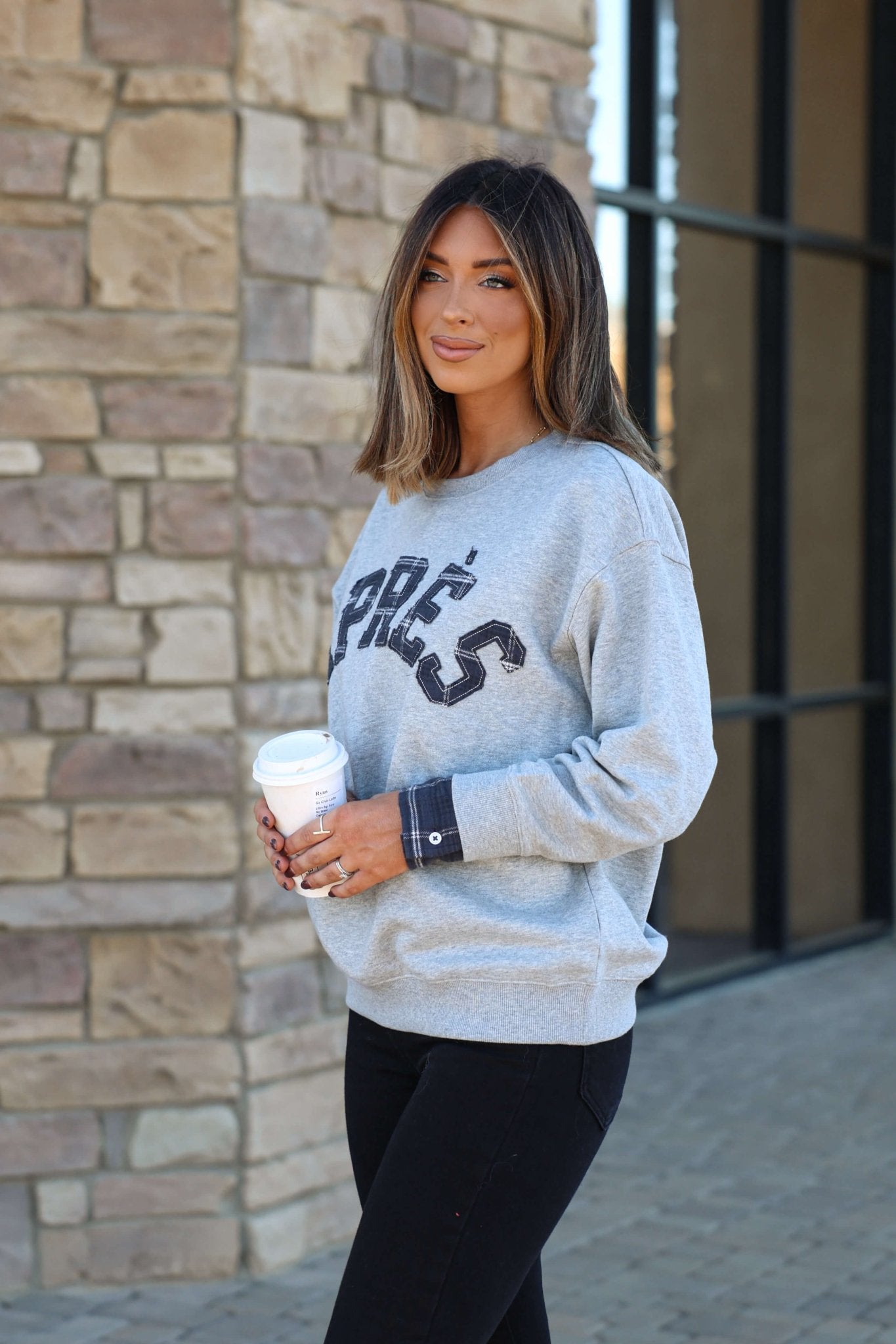 A woman with straight brown hair wears the Vintage Havana Grey Apres French Terry Sweatshirt and holds a coffee cup outside by stone steps.