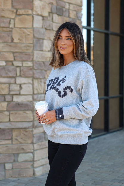A woman with straight brown hair wears the Vintage Havana Grey Apres French Terry Sweatshirt and holds a coffee cup outside by stone steps.