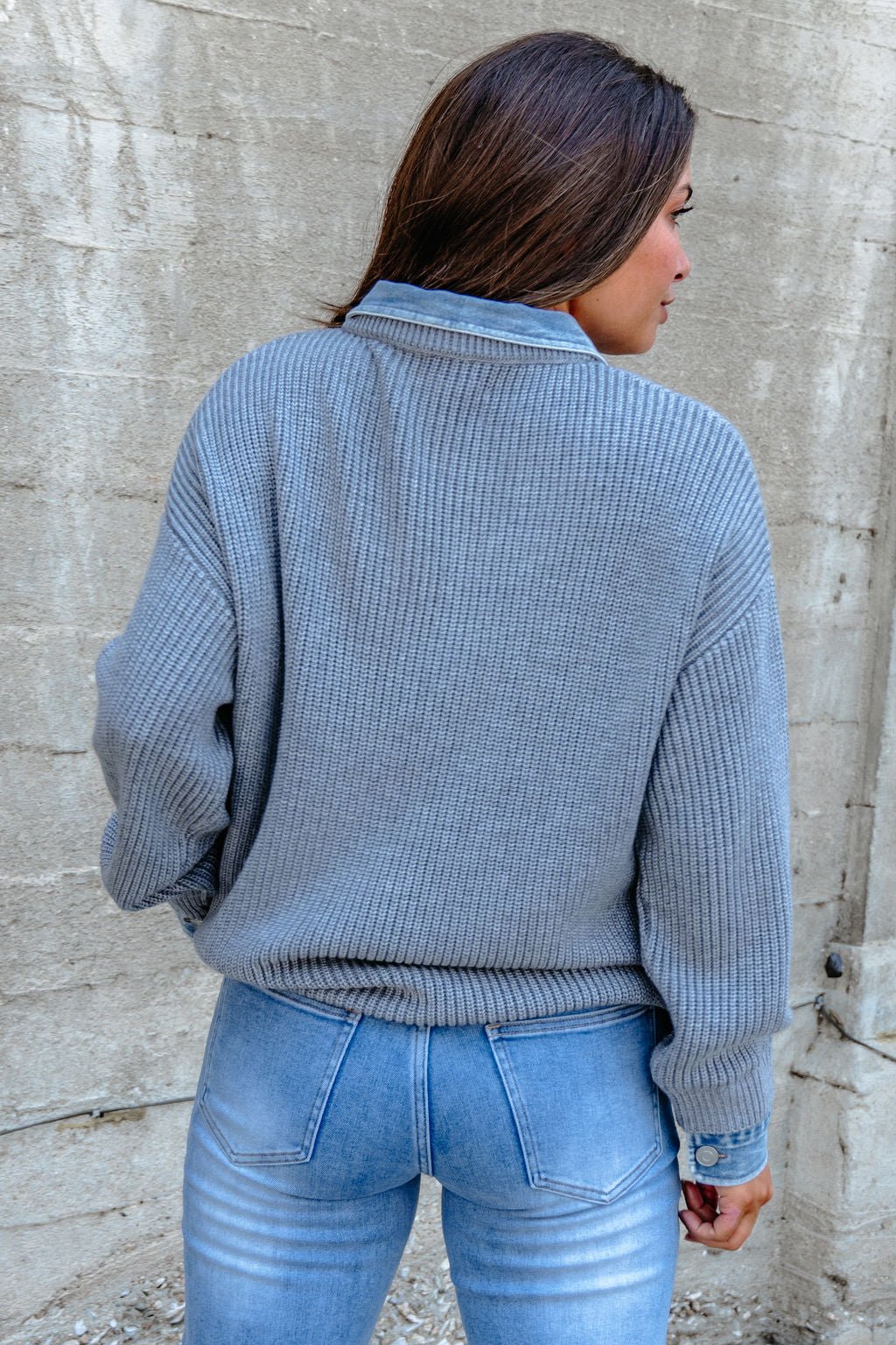 Wearing the Vintage Havana Grey Contrast Ribbed Sweater and light blue jeans, a woman stands with her back to a concrete wall.