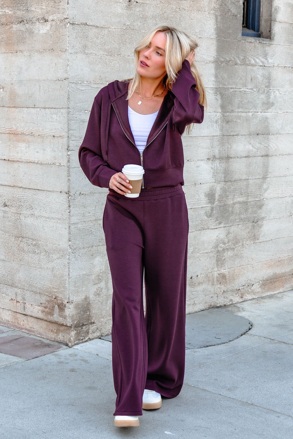 Wearing Vintage Havana Merlot Cloud Fleece Flare Pants, a woman holds coffee and touches her hair by a concrete wall.