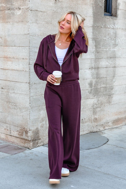 Wearing Vintage Havana Merlot Cloud Fleece Flare Pants, a woman holds coffee and touches her hair by a concrete wall.
