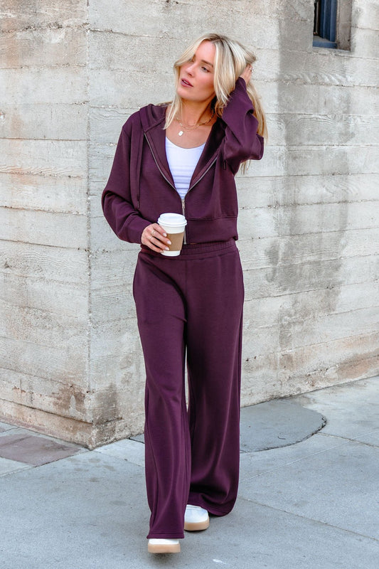 Wearing Vintage Havana Merlot Cloud Fleece Flare Pants, a woman holds coffee and touches her hair by a concrete wall.