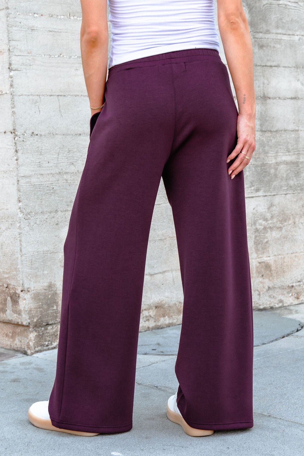 Wearing Vintage Havana Merlot Cloud Fleece Flare Pants, a white top, and white sneakers, standing outdoors by a concrete wall.
