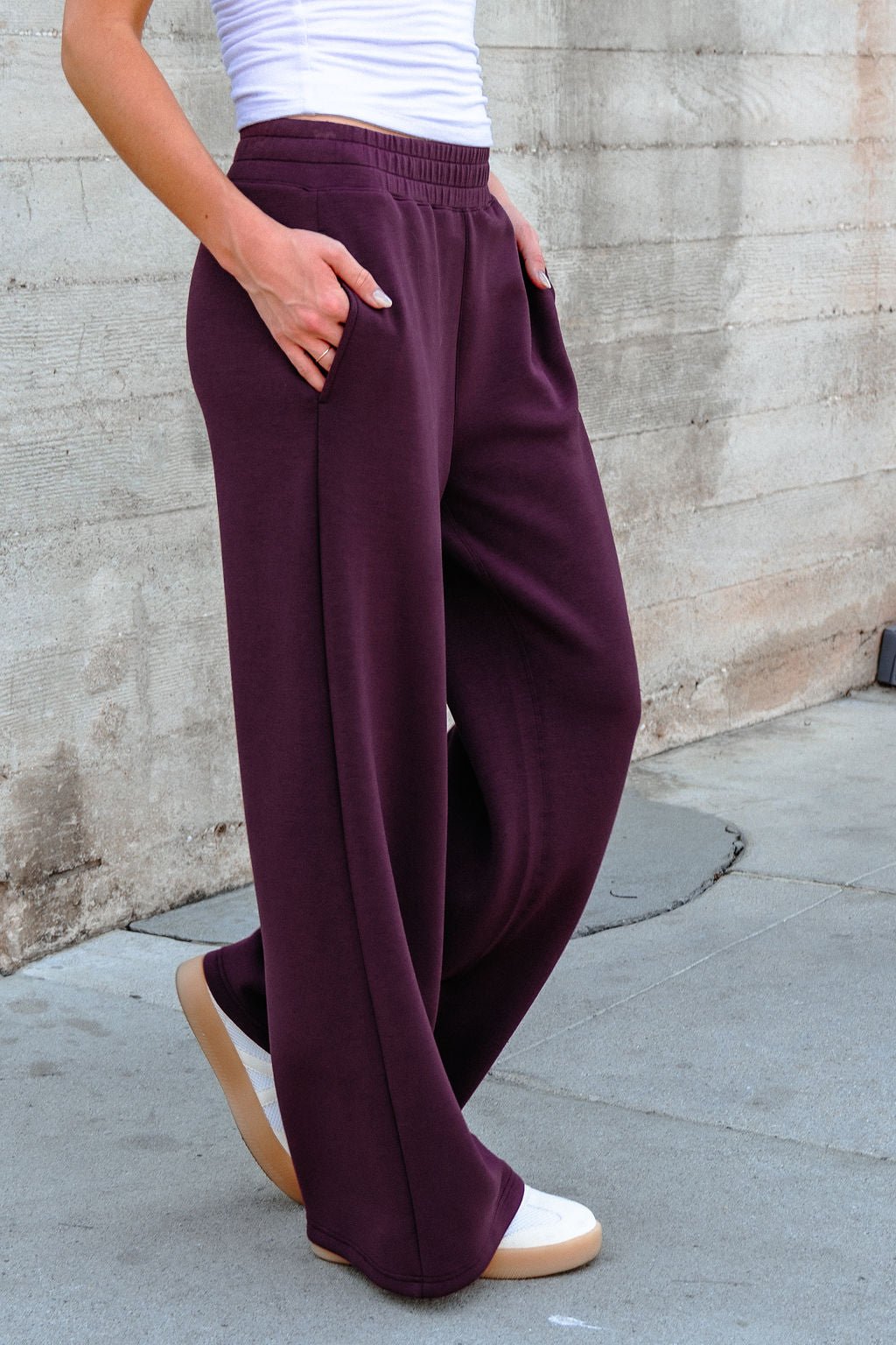 Person wearing Vintage Havana Merlot Cloud Fleece Flare Pants and white shoes, standing against a concrete wall with hands in pockets.